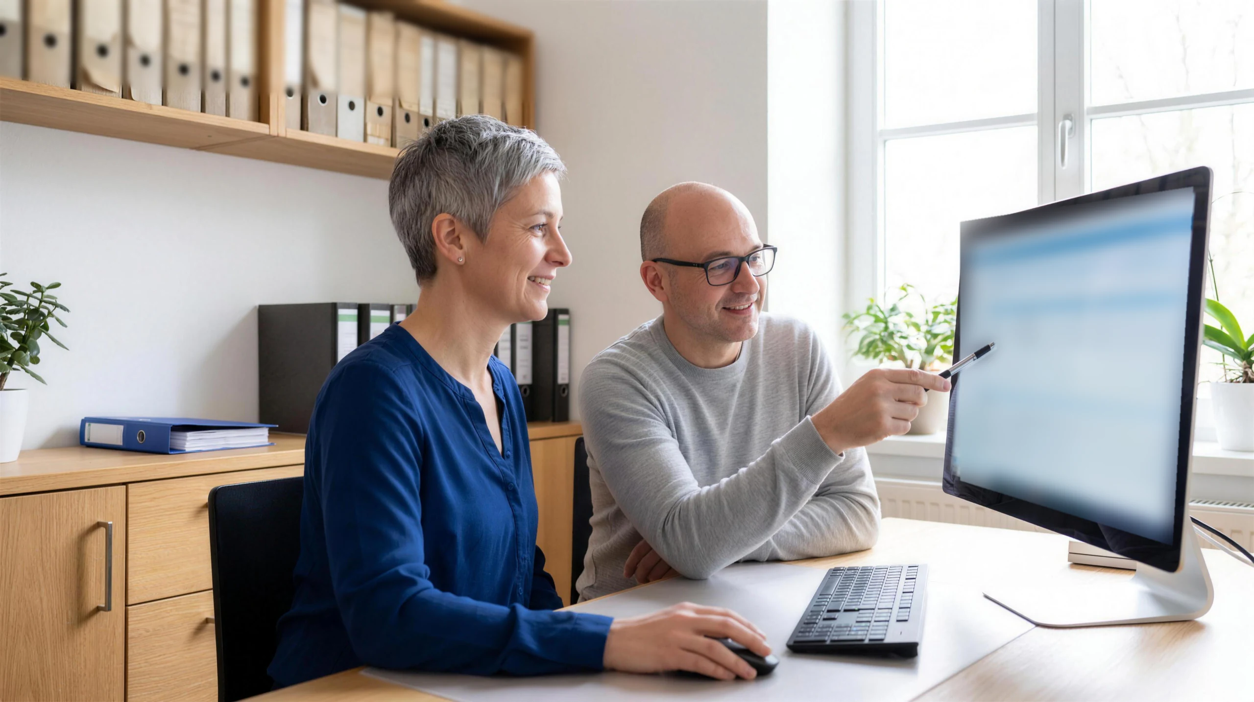 Eine Office-Managerin und ihr Kollege arbeiten am Bildschirm mit ihrer Datenbank, die alten Ordner stehen im Regal